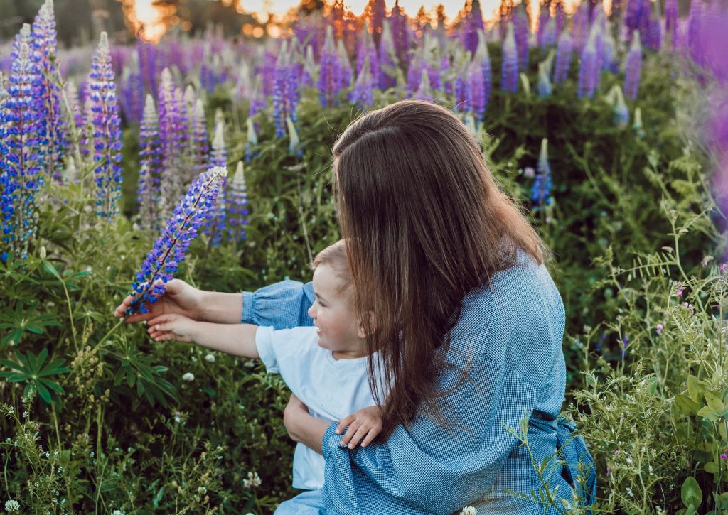 Ung kvinne med langt brunt hår sitter i en eng med et barn på fanget og utforsker de lilla blomstene sammen.