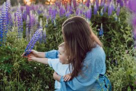 Ung kvinne med langt brunt hår siter på en eng med et barn på fanget og utforsker blomstene sammen.